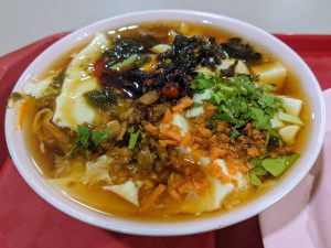 Bowl of northern-style savory doufunao tofu pudding topped with chili paste, cilantro, and preserved vegetables