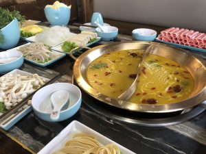 Sichuan-style hotpot table with a bubbling spicy broth pot, plates of raw ingredients, and dipping sauces
