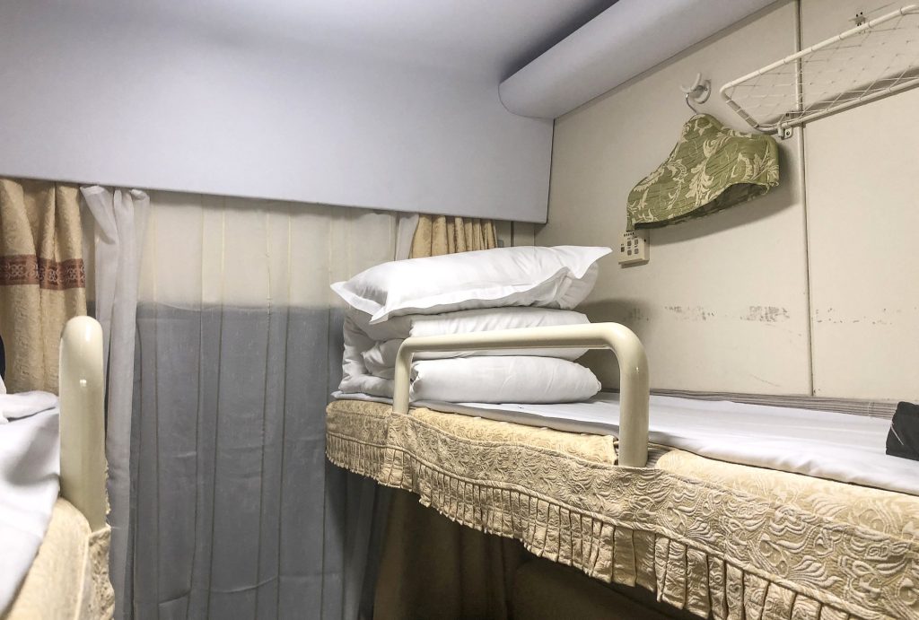 Upper berth inside a China Railway soft sleeper (RW25T) compartment with blue bedding and curtains